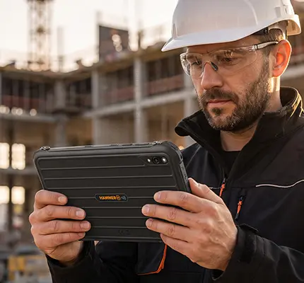 Tablet HAMMER w wytrzymałej czarnej obudowie, używany przez pracownika w kasku i okularach ochronnych na placu budowy