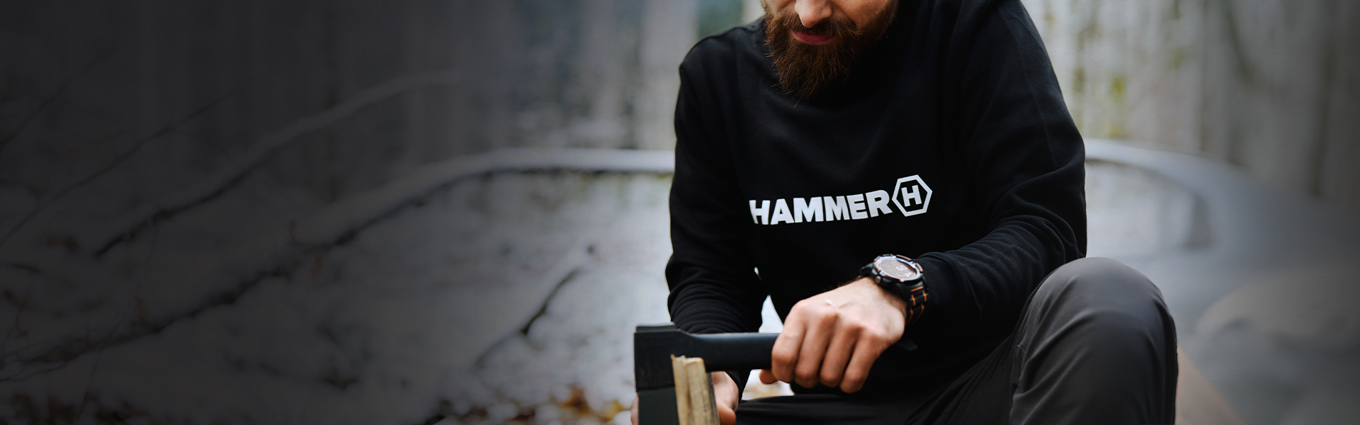 HAMMER CREWNECK mężczyzna siedzi w lesie, siekierą tąbie drzewo. Ma na sobie bluzę  z logiem HAMMER. 