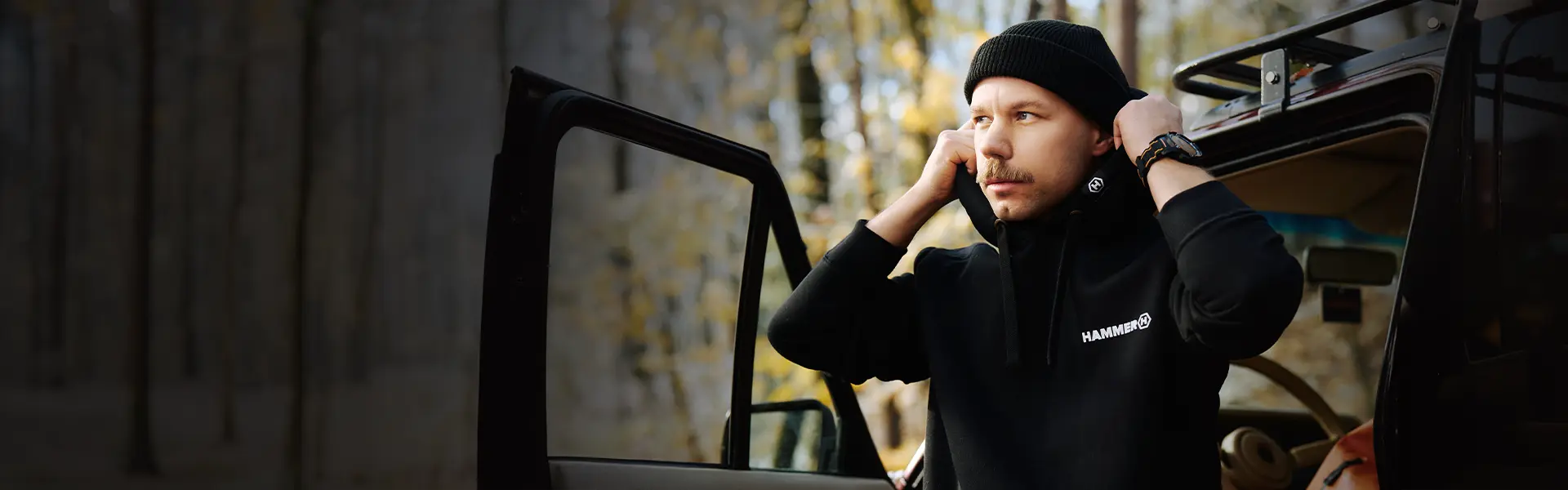 HAMMER HOODIE mężczyzna wysiada z czarnego auta w bluzie HAMMER w lesie.