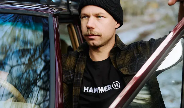 HAMMER T-SHIRT BASIC mężczyzna z wąsem wysiada z bordowego samochodu w lesie.  Ma koszulkę HAMMER,  na niej koszulę w kratę. 