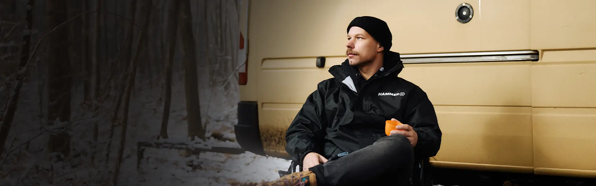 HAMMER JACKET mężczyzna podczas campingu zimą , siedzi na krześle turystycznym obok żółtego busa.  Ma na sobie czarną kurtkę HAMMER.