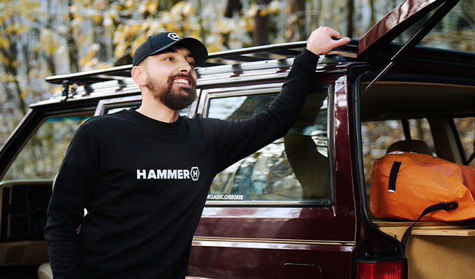 A man is leaning against a car, wearing a HAMMER WEAR cap and sweatshirt.