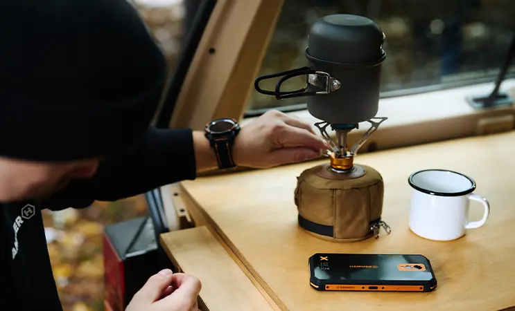HAMMER na campingu, stolik, a na nim telefon wzmocniony HAMMER , metalowy kubek i kuchenka turystyczna